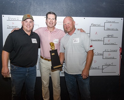 Three men holding trophy standing in front of Cornhole tournament bracket